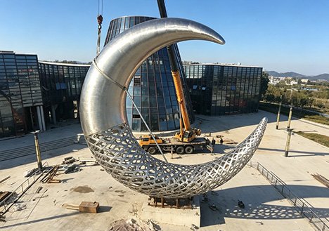 Monumental moon-shaped Custom Stainless Steel Sculptures being installed with a crane at commercial complexes, featuring FEA-validated structural integrity for office campuses.