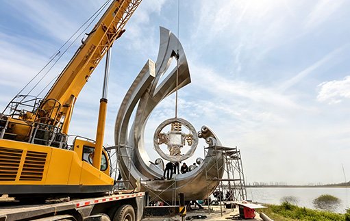 Large yellow crane performing Crane Rigging & Lifting Support for a monumental stainless steel sculpture, demonstrating Wensculp's Technical Support for Custom Sculpture Installation and Unpacking & Handling Protocols for large-scale projects.